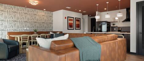 A modern living room with a brown leather sofa, a dining area with a table and chairs, and a kitchen with stainless steel appliances.
