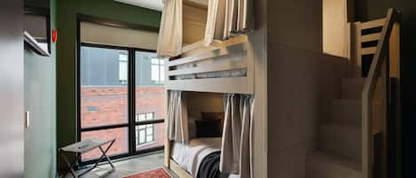 Una habitación con una cama litera, una mesita, una silla, un ventanal con vista a un edificio de ladrillo y una escalera.
