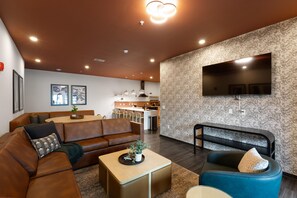 A modern living room with a brown leather sofa, a blue armchair, and a coffee table. There is a kitchen in the background with a large TV mounted on the wall. - Superior Suite - Unit 702 (Nashville)