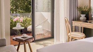A hotel room with a bed, a desk with a chair, a small table with a vase and flowers, and a sliding glass door leading to an outdoor balcony with a chair and plants. - LoverA Signature Hotel (Ho Chi Minh City)