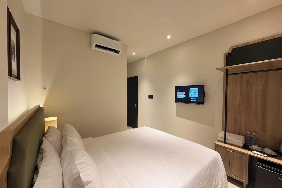 A hotel room with a bed, a wall-mounted TV, a wardrobe, and a small table with a lamp.