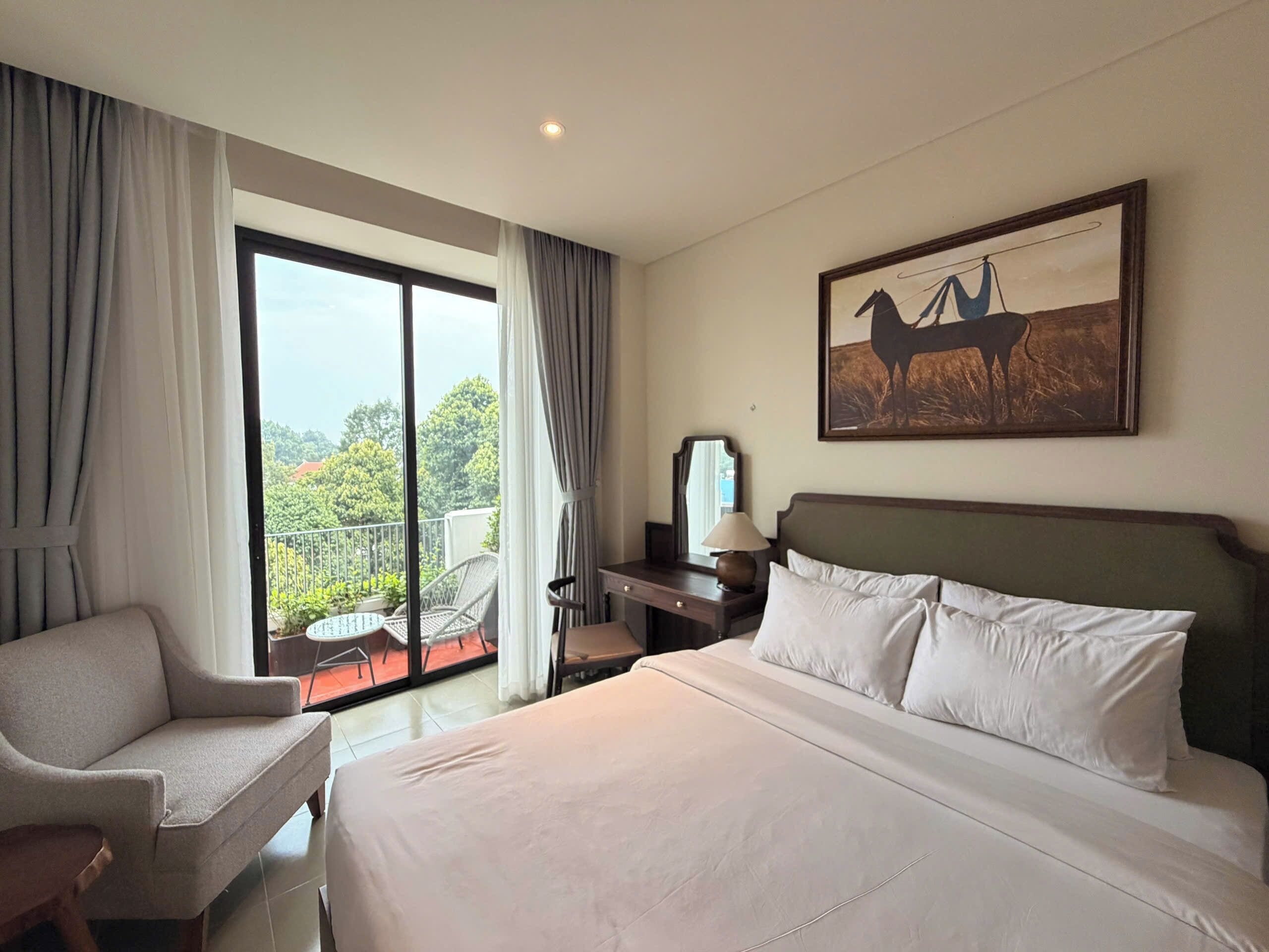 A hotel room with a bed, a chair, a desk, a lamp, a painting, and a balcony with a view.