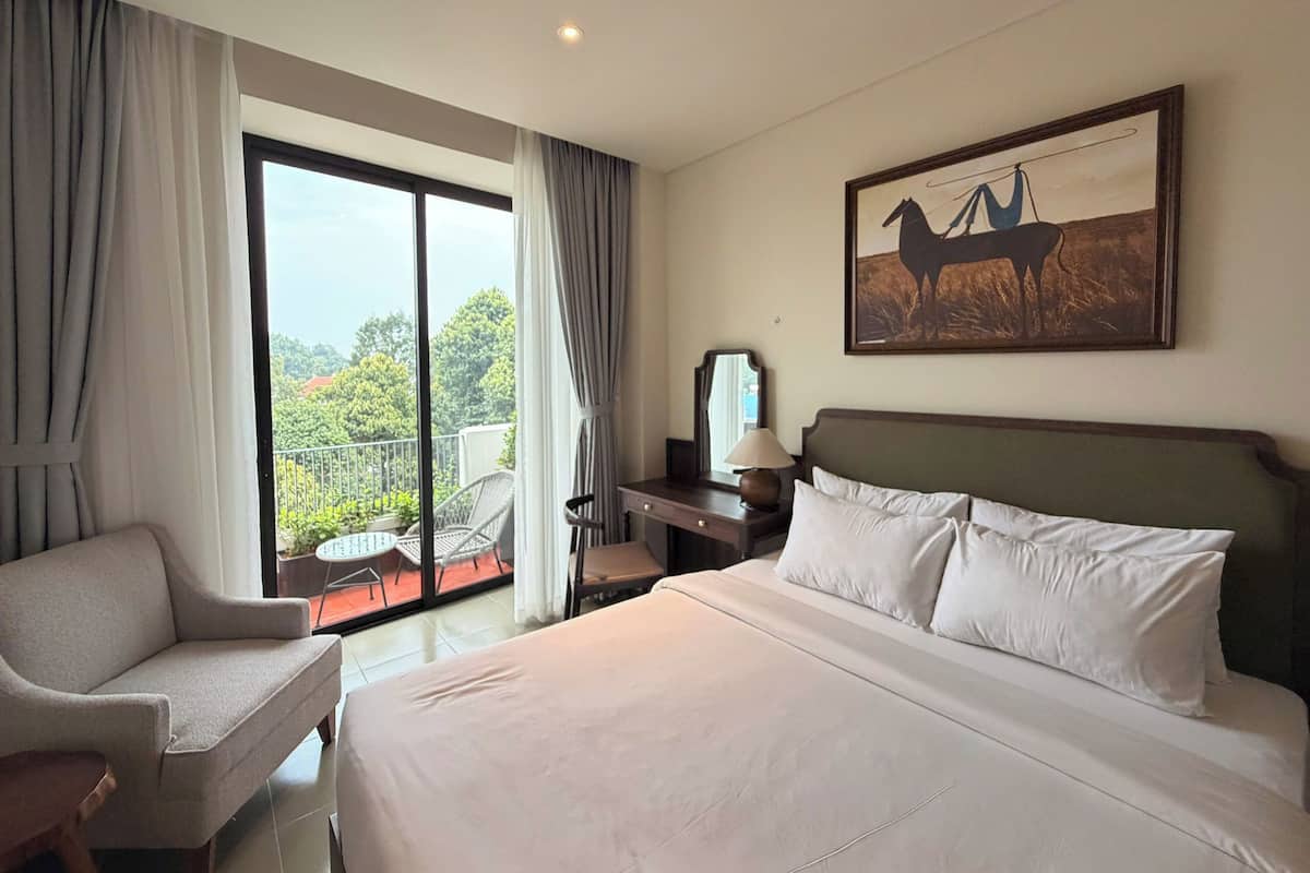 Habitación de hotel con cama, una silla, escritorio, lámpara, un cuadro y balcón con vistas.