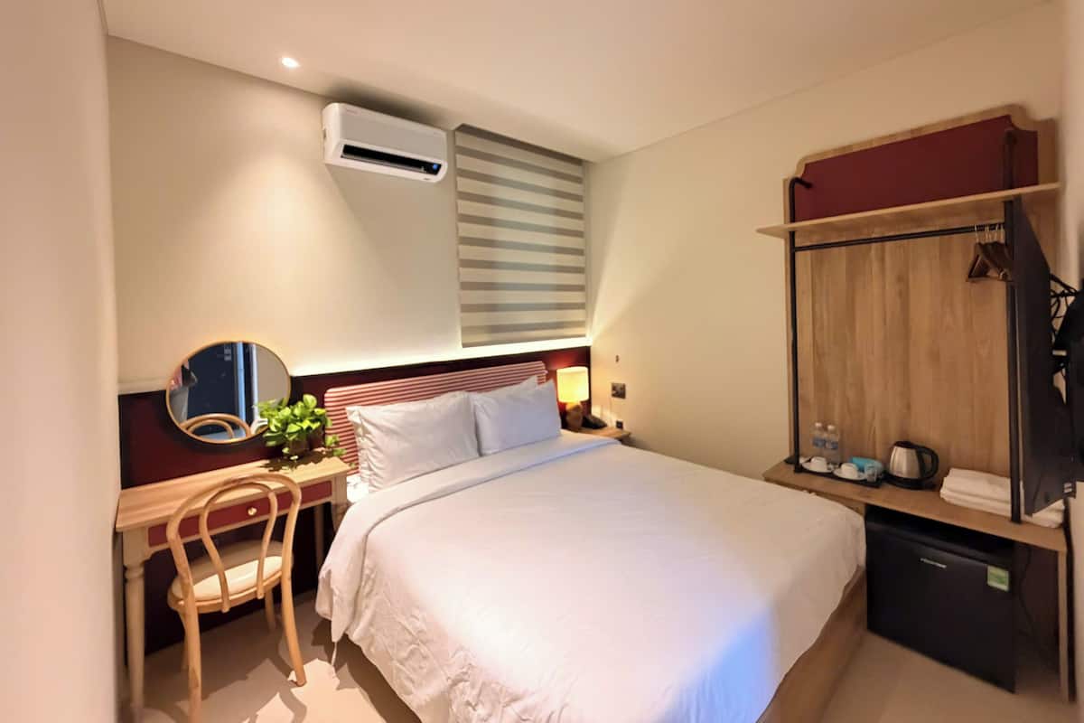 A hotel room with a bed, a desk with a chair, a mirror, a bedside table with a lamp, a wardrobe, and an air conditioning unit.