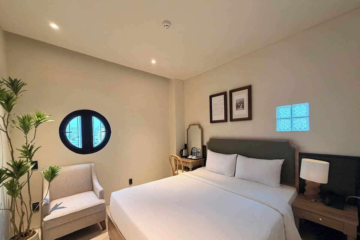 A bedroom with a bed, a chair, a nightstand, a lamp, a mirror, a window, and a plant.