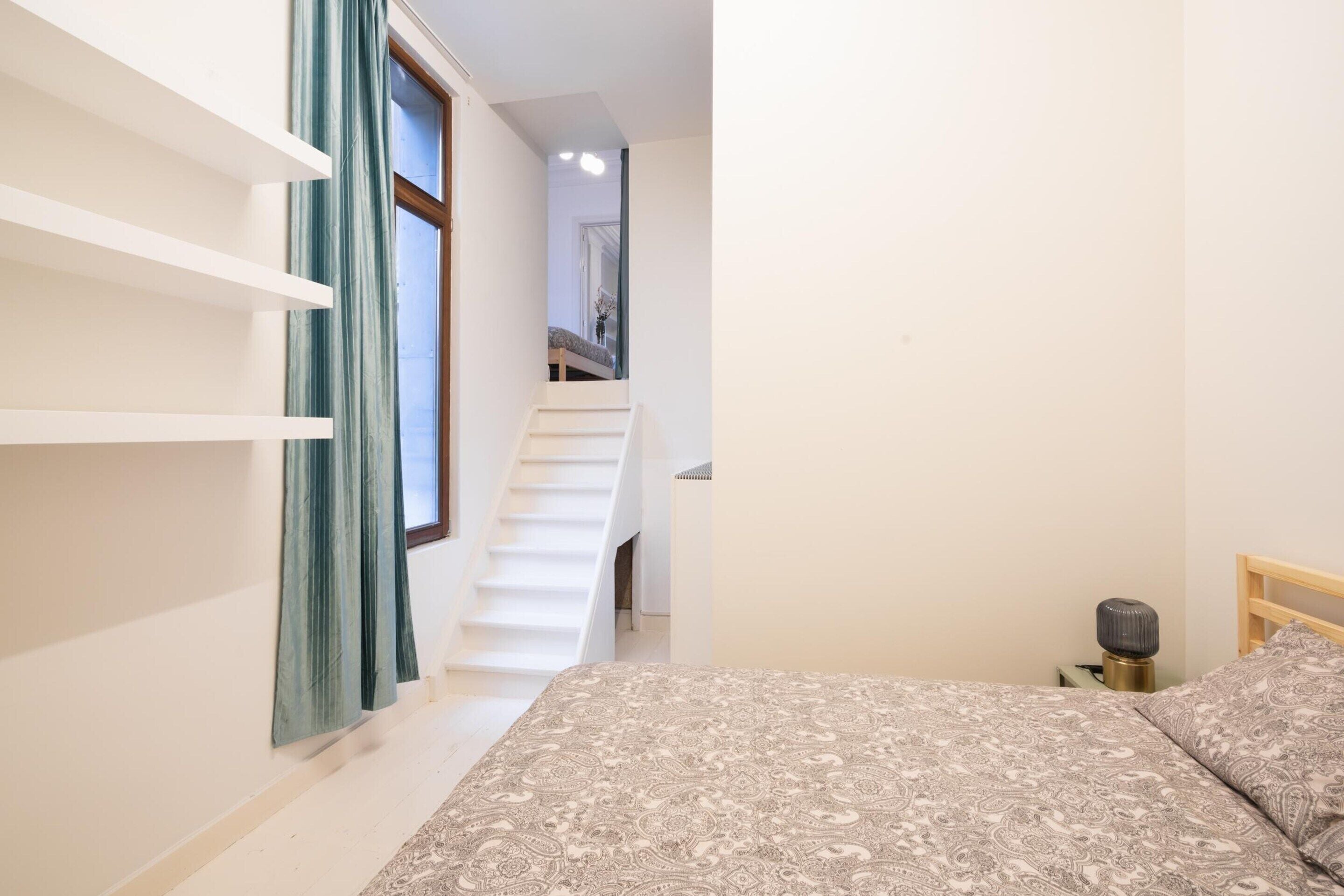 A bedroom with a bed, a bedside table, a lamp, a window with curtains, and a staircase leading to another room.
