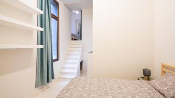 A bedroom with a bed, a bedside table, a lamp, a window with curtains, and a staircase leading to another room.