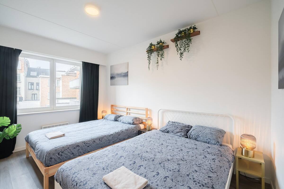 Habitación de hotel con dos camas, ventana, radiador, planta en la pared y lámpara de noche.