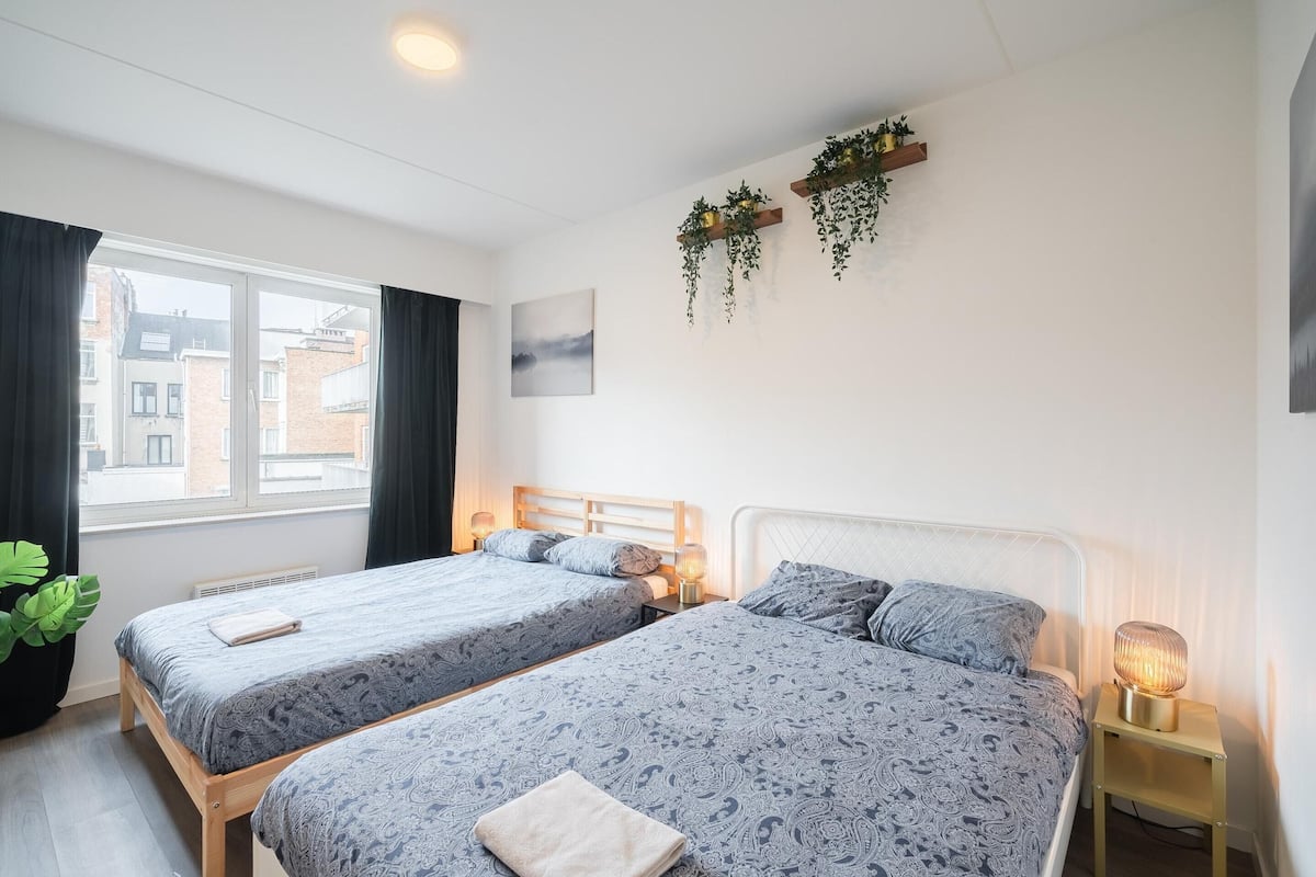 Ett hotellrum med två sängar, ett fönster, ett element, en växt som hänger på väggen och en sänglampa.