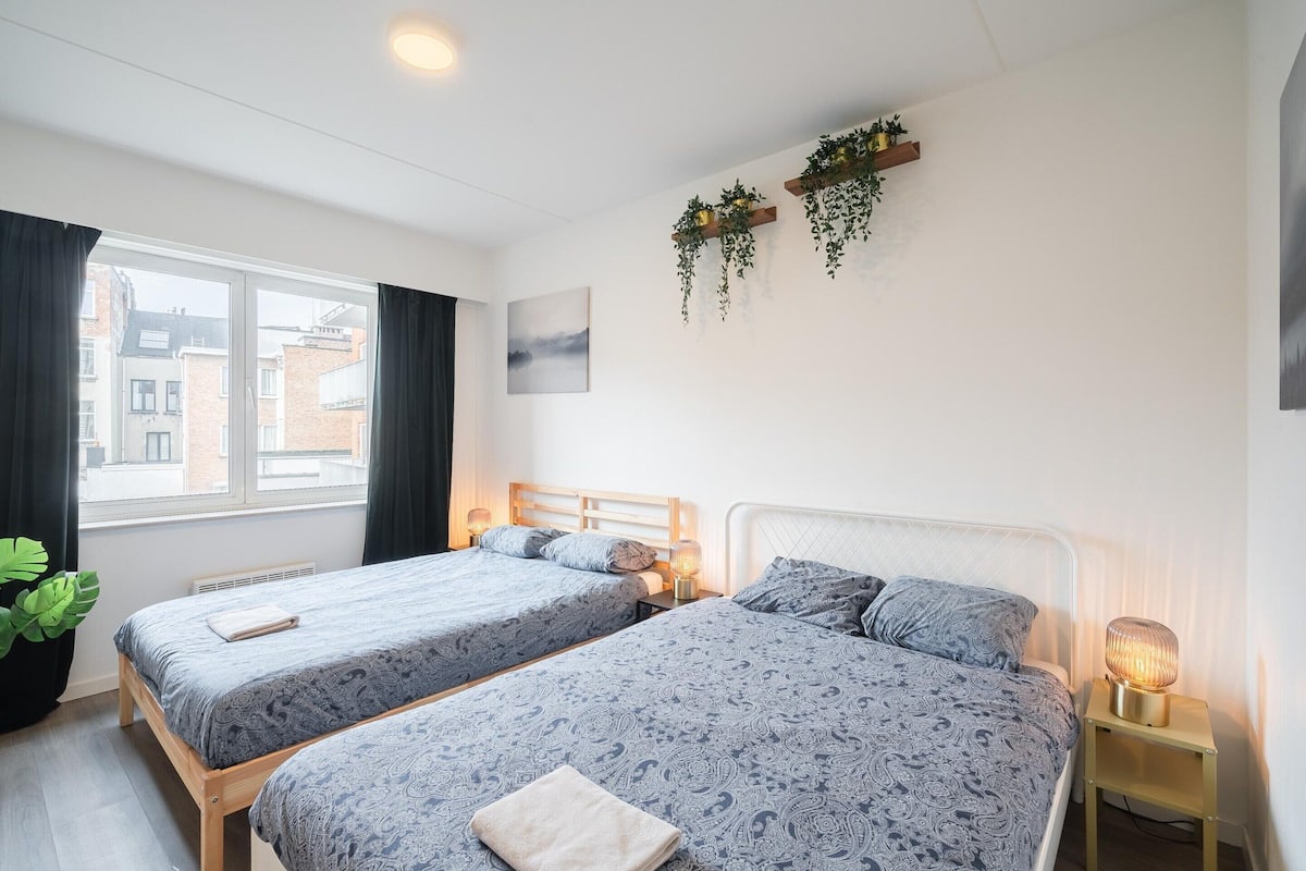 Habitación de hotel con dos camas, una ventana con cortinas, una planta en la pared y una lámpara de noche.