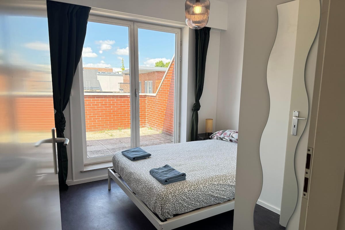Un dormitorio con cama, ventana con cortinas, una puerta y vista a un edificio de ladrillo.