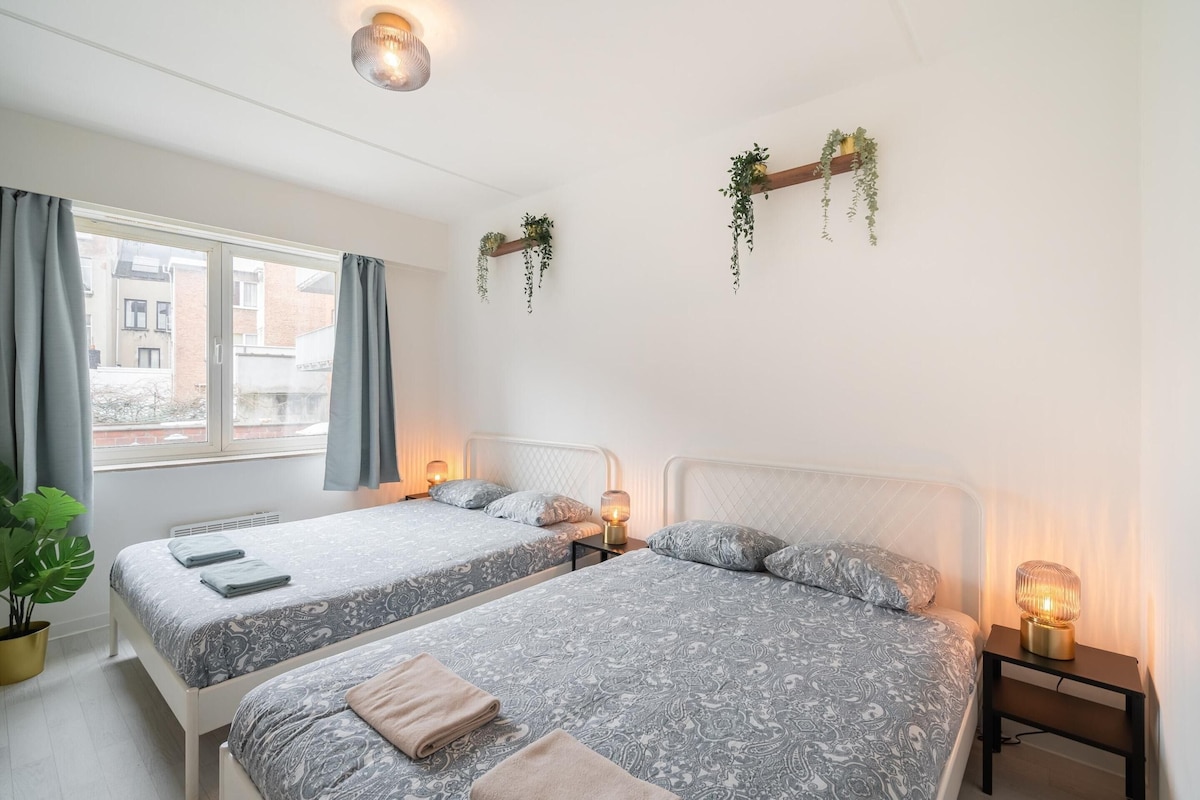 A room with two beds, a large window, and hanging plants.
