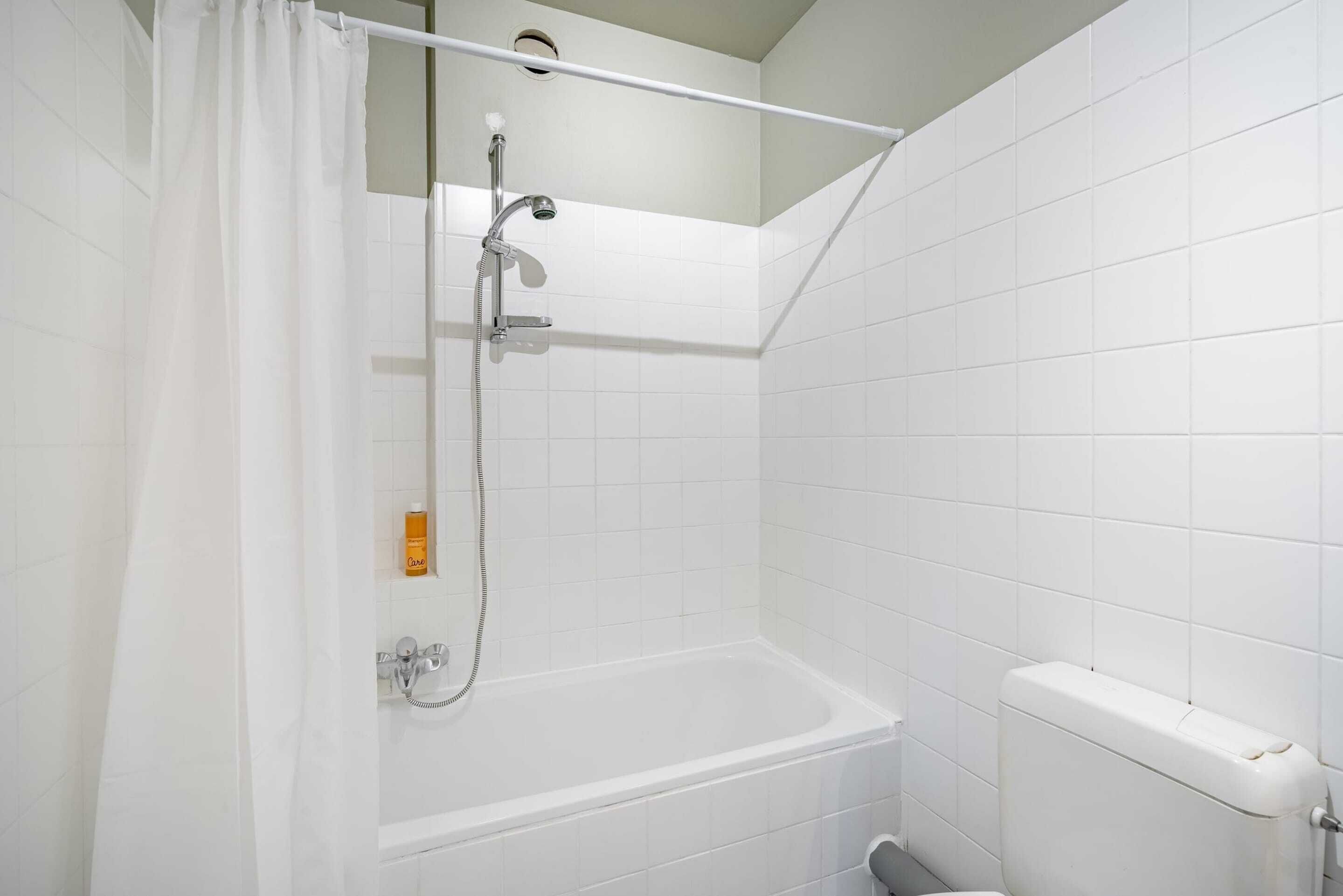 Une salle de bain avec des murs recouverts de carreaux blancs, une douche avec un rideau, une baignoire et des toilettes.