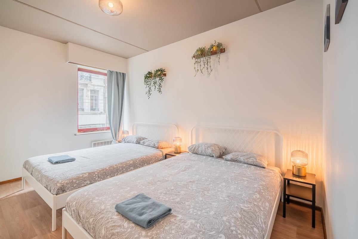 Un dormitorio con dos camas, una ventana con cortinas, una mesita de noche con lámpara y macetas colgantes en la pared.