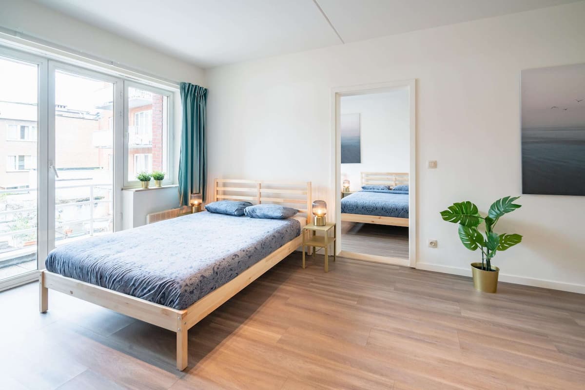 Un dormitorio con una cama de madera, una mesita de noche con lámpara, una planta en maceta y un ventanal grande.