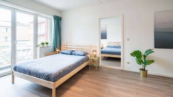 A bedroom with a wooden bed, a bedside table with a lamp, a potted plant, and a large window.