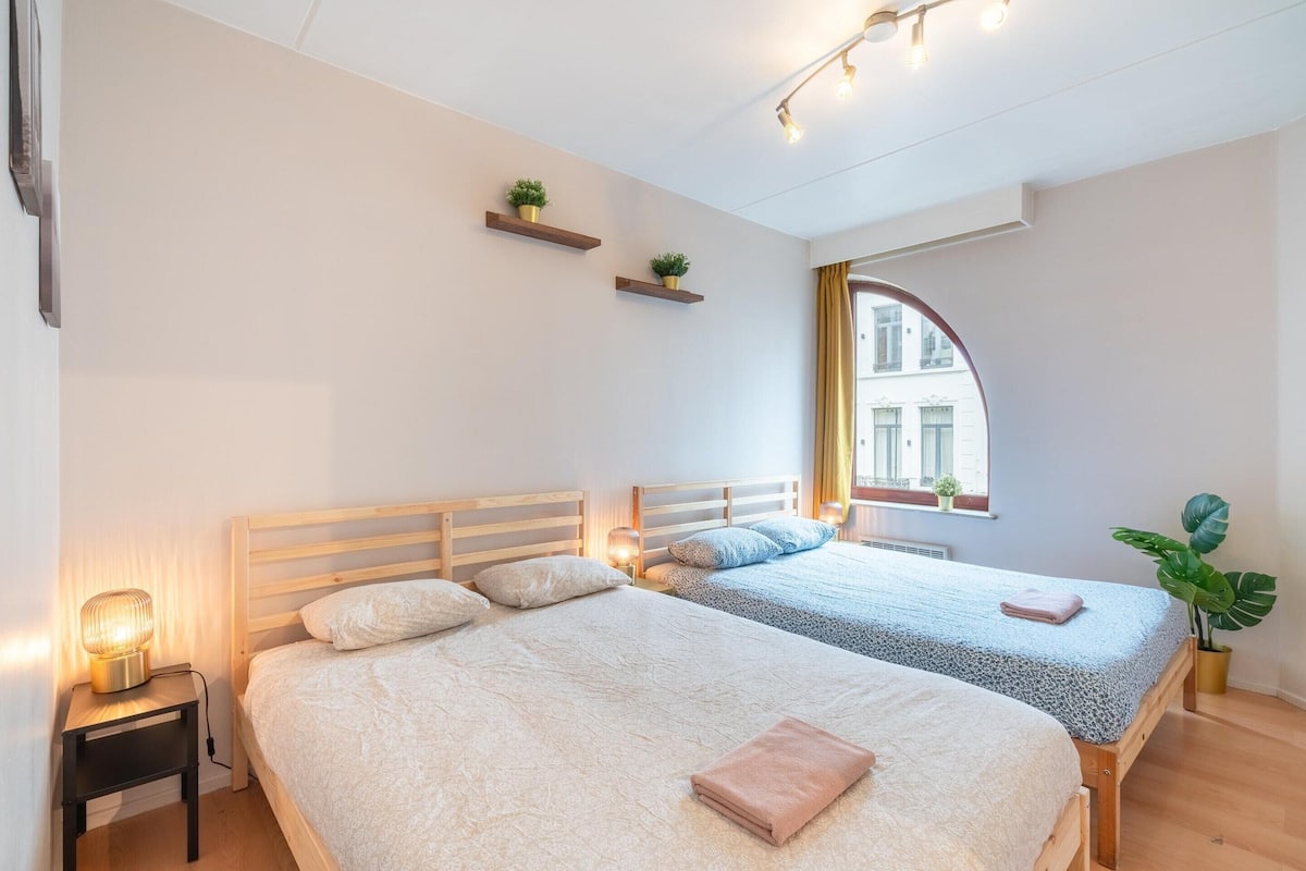 Un dormitorio con dos camas, mesitas de noche, una ventana con cortinas y una planta en maceta.