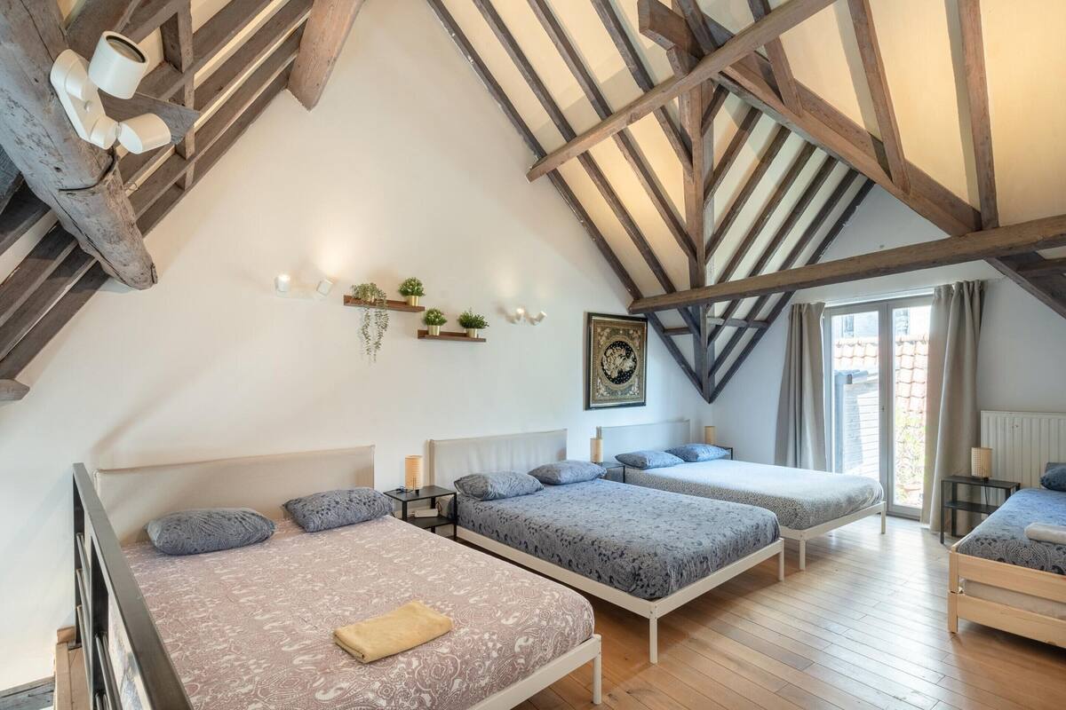 A loft-style bedroom with exposed wooden beams, three beds, a large window, and a view of the outdoors.
