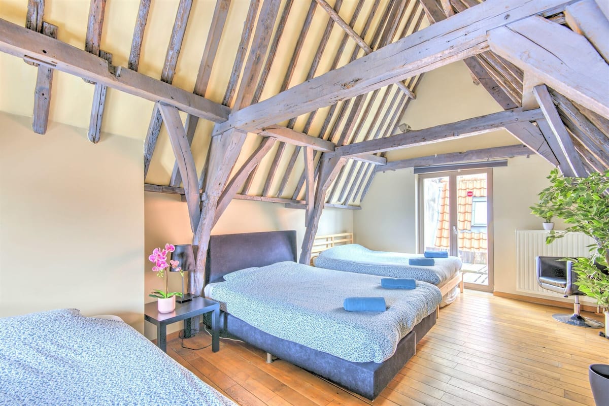 A room with a sloped ceiling, wooden beams, two beds with blue bedding, a small table with a lamp, and a chair.