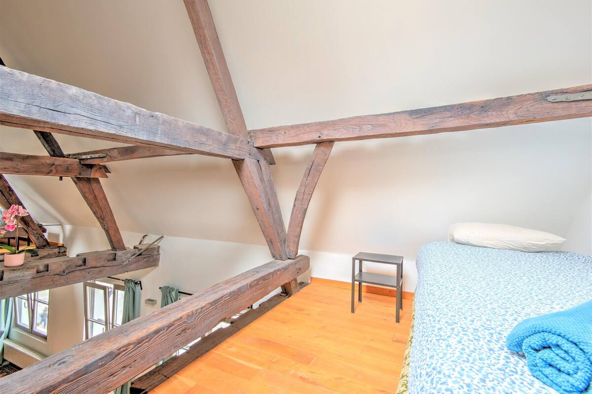 A room with exposed wooden beams, a bed with a patterned bedspread, a small table, and a window with curtains.