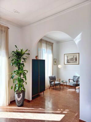 Una habitación con una planta grande en maceta, un armario de color oscuro, dos sillas estampadas, una mesita y un cuadro en la pared.