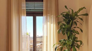 Ein Zimmer mit Schiebeglastür, beigen Vorhängen, einer großen Kübelpflanze und Blick auf einen Balkon mit gelben Geländern.