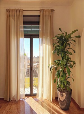 Una habitación con puerta de vidrio corredera, cortinas color beige, una planta grande en maceta y vistas a un balcón con barandillas amarillas.