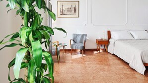 Ein Zimmer mit einem Bett, einem Sessel, einem kleinen Tisch, einer Pflanze und einem gerahmten Bild an der Wand.