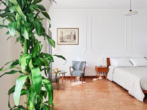 Una habitación con una cama, una silla, una mesita, una planta y un cuadro en la pared.