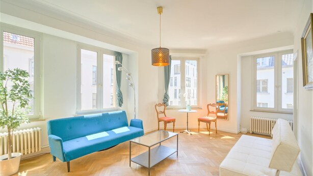 Een woonkamer met een blauwe bank, een witte bank, een glazen salontafel en een houten stoel.