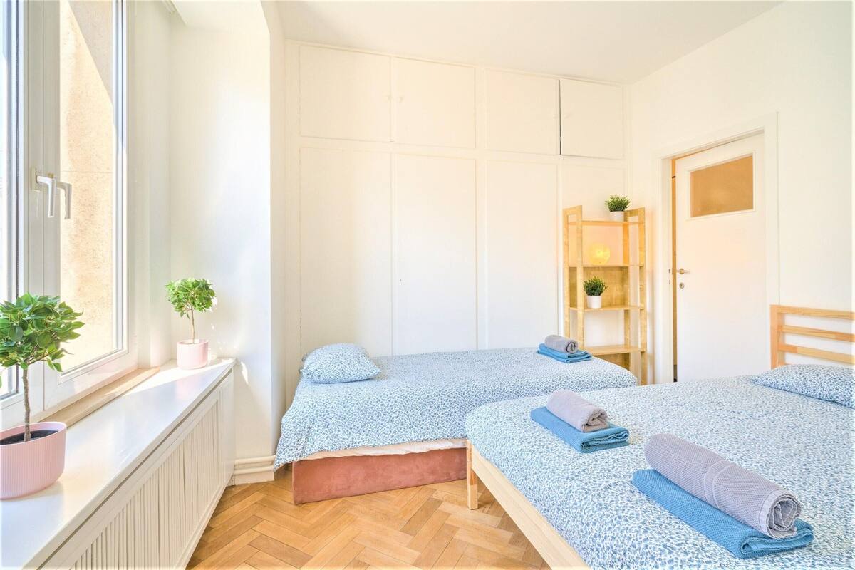 Une chambre avec deux lits, un radiateur, une fenêtre ornée de plantes et une porte en bois.
