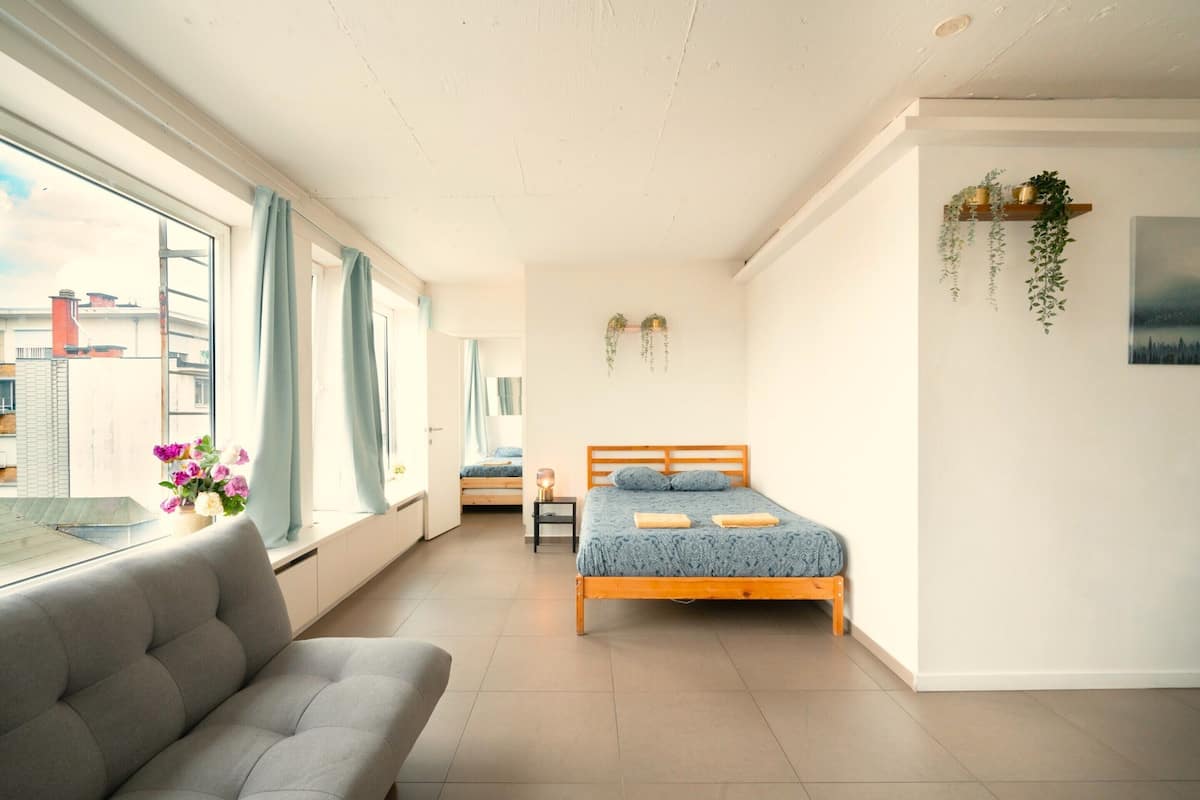 A bedroom with a bed, a sofa, a window with curtains, a small table, and a wall-mounted shelf with plants.