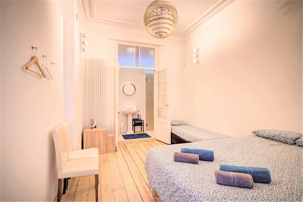 A bedroom with a bed, a chair, a small table, a radiator, and a door leading to another room.