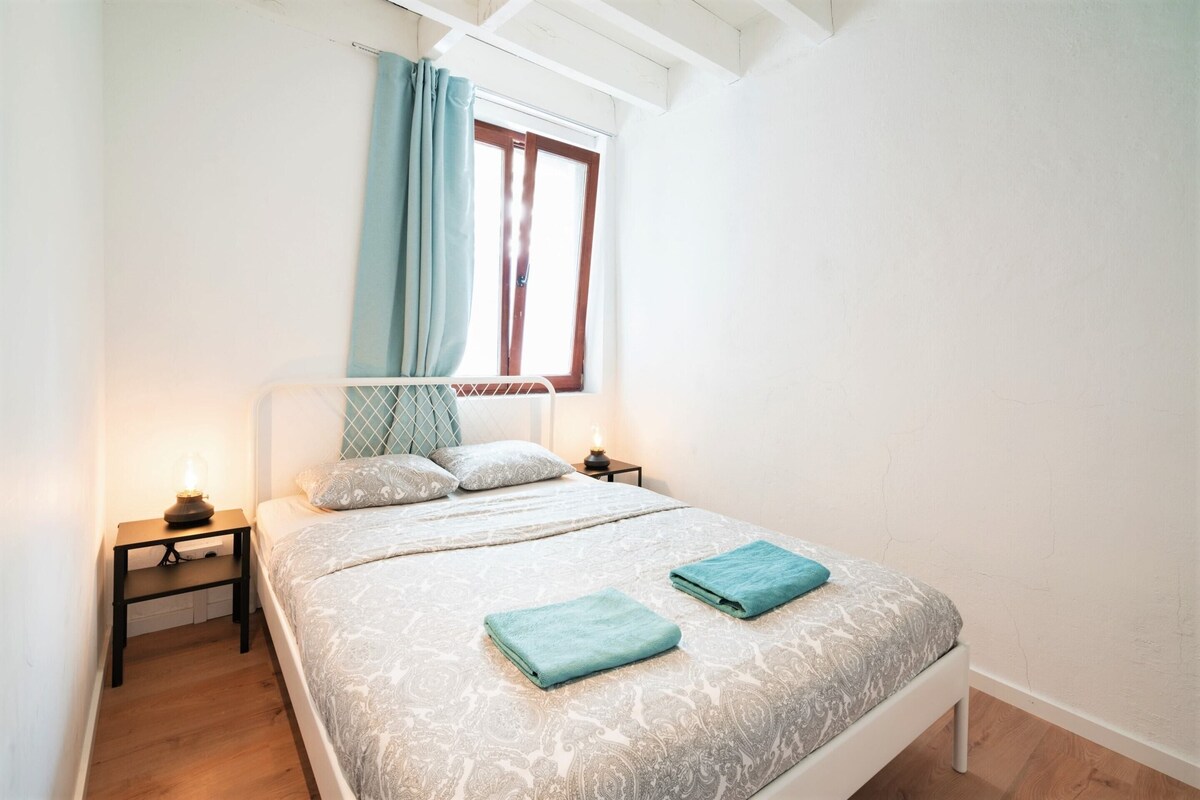 A bedroom with a bed, a nightstand, a window with curtains, and a bedside lamp.