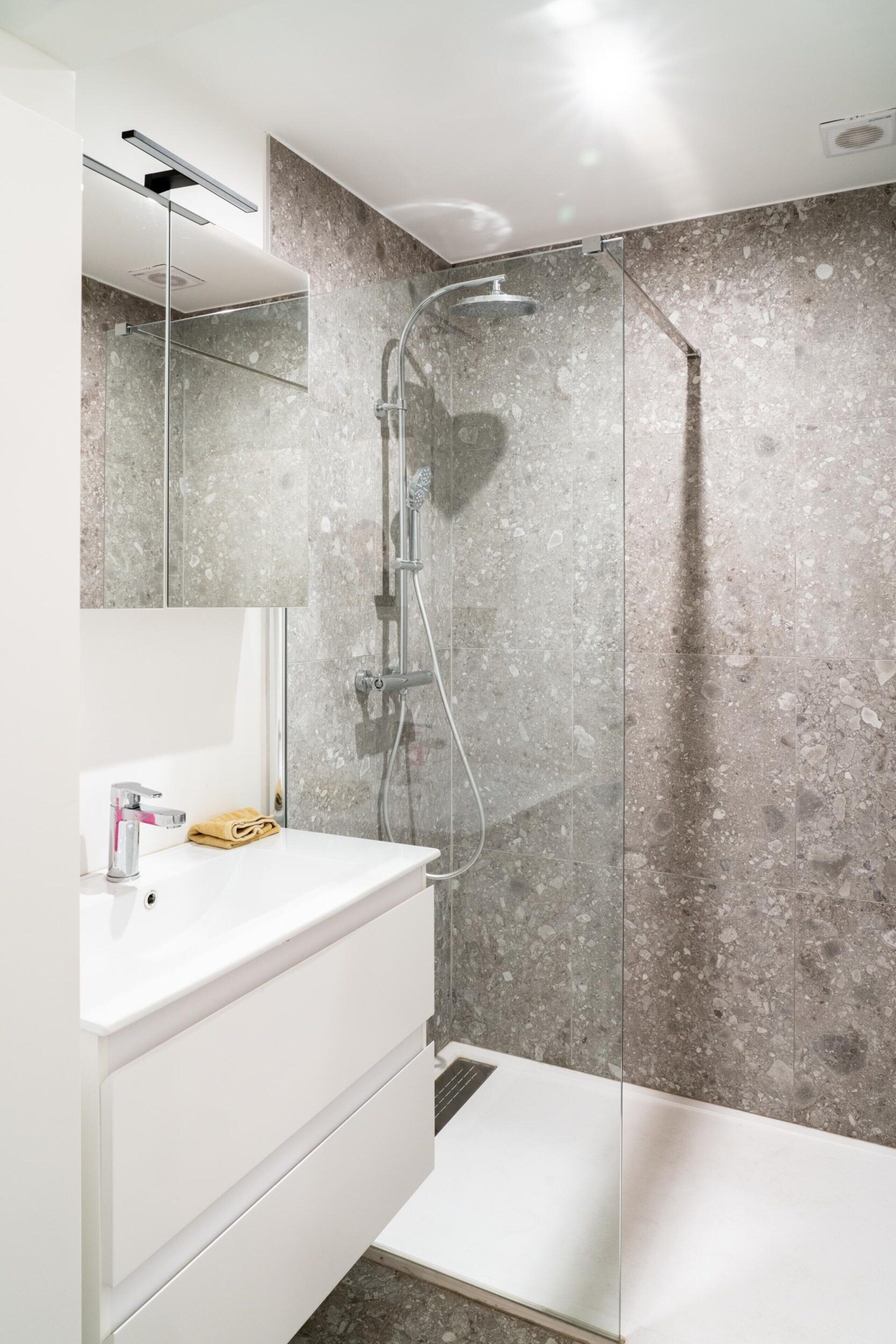 Un baño moderno con ducha acristalada, tocador blanco con espejo y salpicadero de mármol.