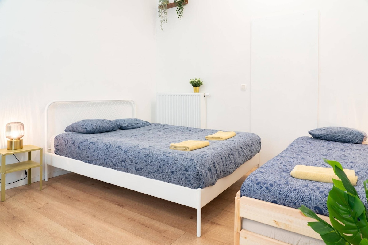 A room with two beds, a bedside table with a lamp, a radiator, and a small plant.
