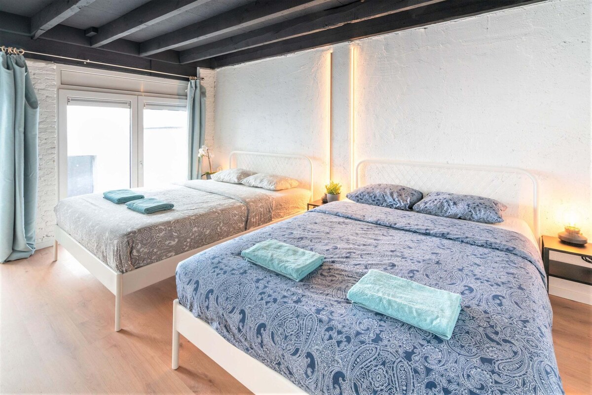 A bedroom with two beds, a window with curtains, and a nightstand with a lamp.