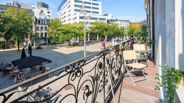A balcony with a wrought iron railing, outdoor seating, and a view of a city street with buildings and trees.