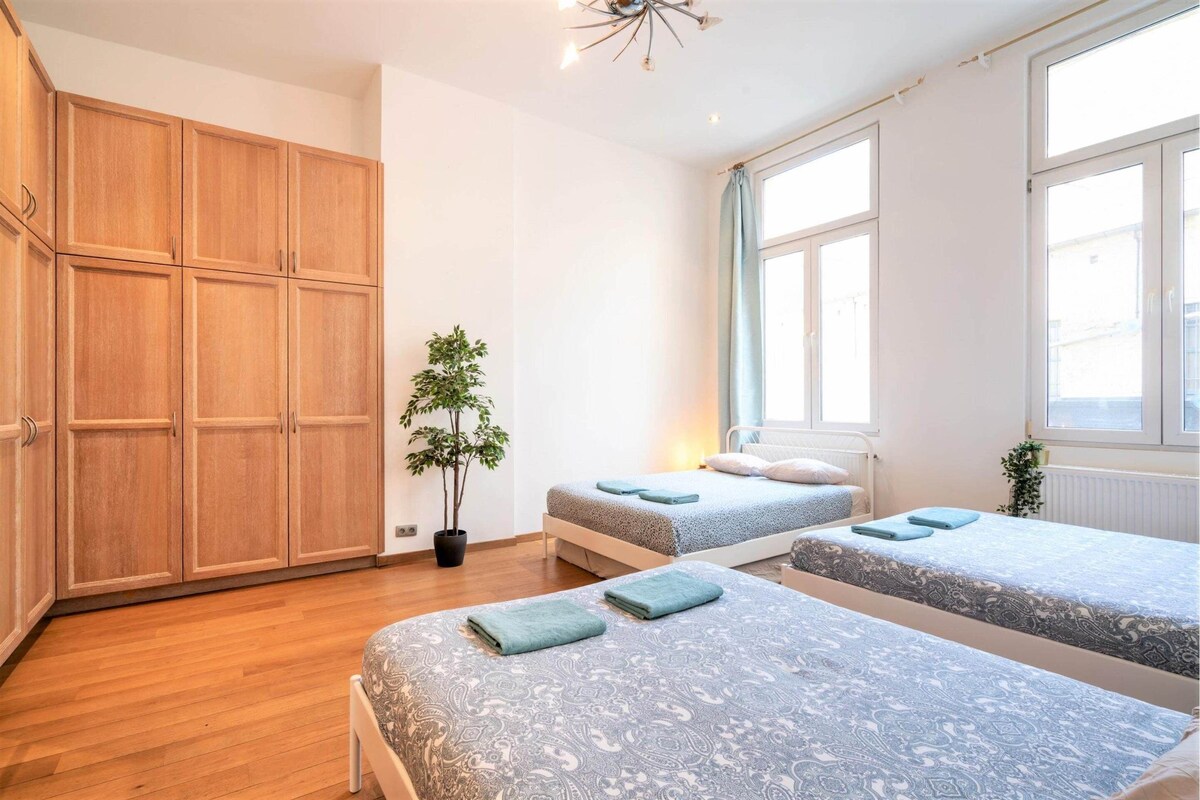 A bedroom with two beds, wooden wardrobe, potted plant, and large windows.