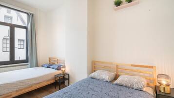 A bedroom with two beds, a window, a bedside table with a lamp, and a small shelf with plants.