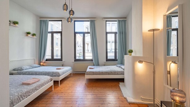 Quarto com três camas, piso de madeira, janelas amplas com cortinas, abajur e espelho.
