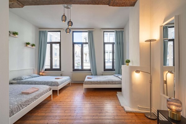 A room with three beds, wooden flooring, large windows with curtains, a standing lamp, and a mirror. - Lively City Getaway for 10 Near Meir (Antwerp)