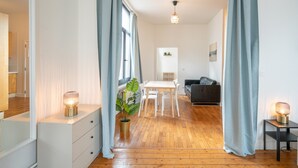 Quarto com piso de madeira, mesa branca com cadeiras, sofá de couro preto, planta em vaso e pequena mesa com abajur.