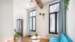 Quarto com sofá azul, piso de madeira, mesa redonda e janelas amplas com cortinas.