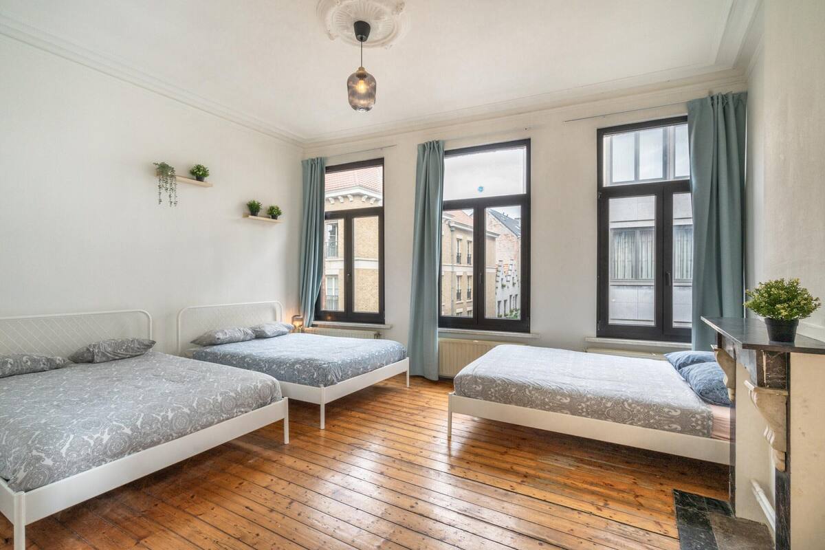 A room with three beds, wooden flooring, and large windows with curtains.
