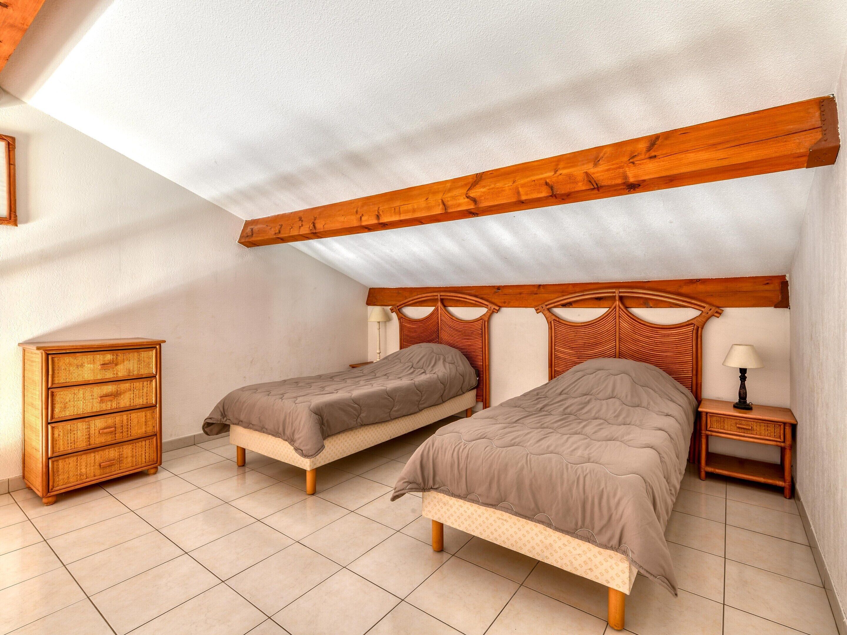 A bedroom with two single beds, a wooden headboard, a nightstand, a dresser, and a tiled floor.