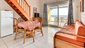 A room with a dining table, chairs, a sofa, a wooden staircase, a refrigerator, a television, and a view of the sea through a sliding glass door. - Large Seaview Stay for 8 in Valras Plage (Valras-Plage)