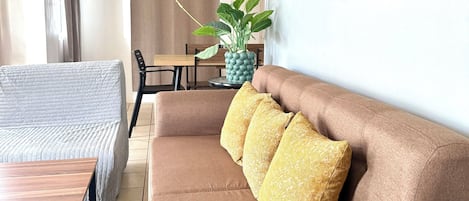A living room with a brown sofa, a wooden coffee table, a grey armchair, a dining table with chairs, and a potted plant.