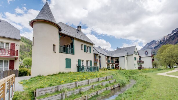 A traditional chalet-style building with a prominent tower, a small stream, and a mountainous backdrop. - 4-guest Studio Near Thermal Baths (Loudenvielle)