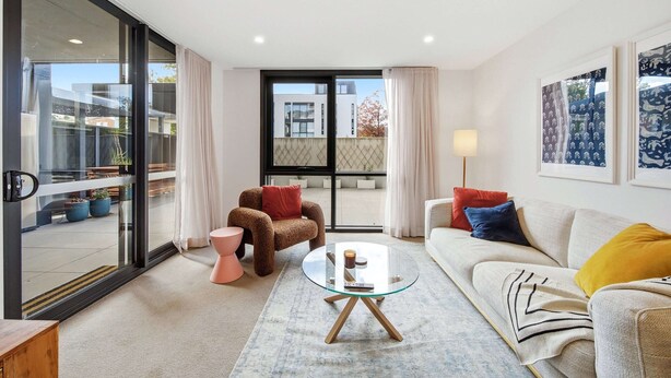Ein modernes Wohnzimmer mit einem Glastisch, einer Couch, einem Sessel und Blick durch eine Glastür auf einen Balkon.